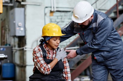 Image is of a male worker with hard hat helping female worker with a hurt shoulder concept of preventing workplace injuries