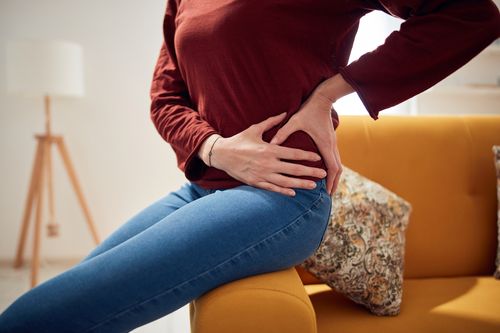 Image is of a woman sitting on her couch with her hands on her hip indicating hip pain, concept of hip pain treatment in Chicago
