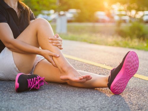 Image is of a female runner sitting on the ground holding her  ankle which is in pain, concept of muscle sprain and strain treatment in Chicago