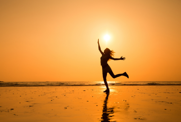 Image is of a happy woman dancing on the beach, concept of how massage therapy can help with reduction of pain
