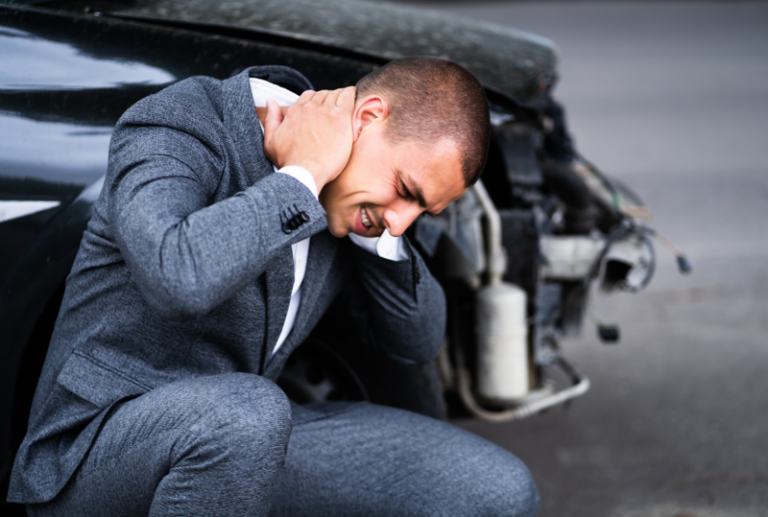 Image is of a man kneeling next to his wrecked car and holding his neck. Concept of whiplash injury and treatment