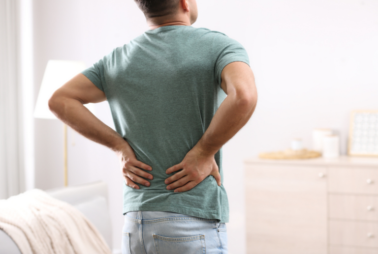 Image is of a man in pain standing with his hands on his lower back, concept of back pain after car accident